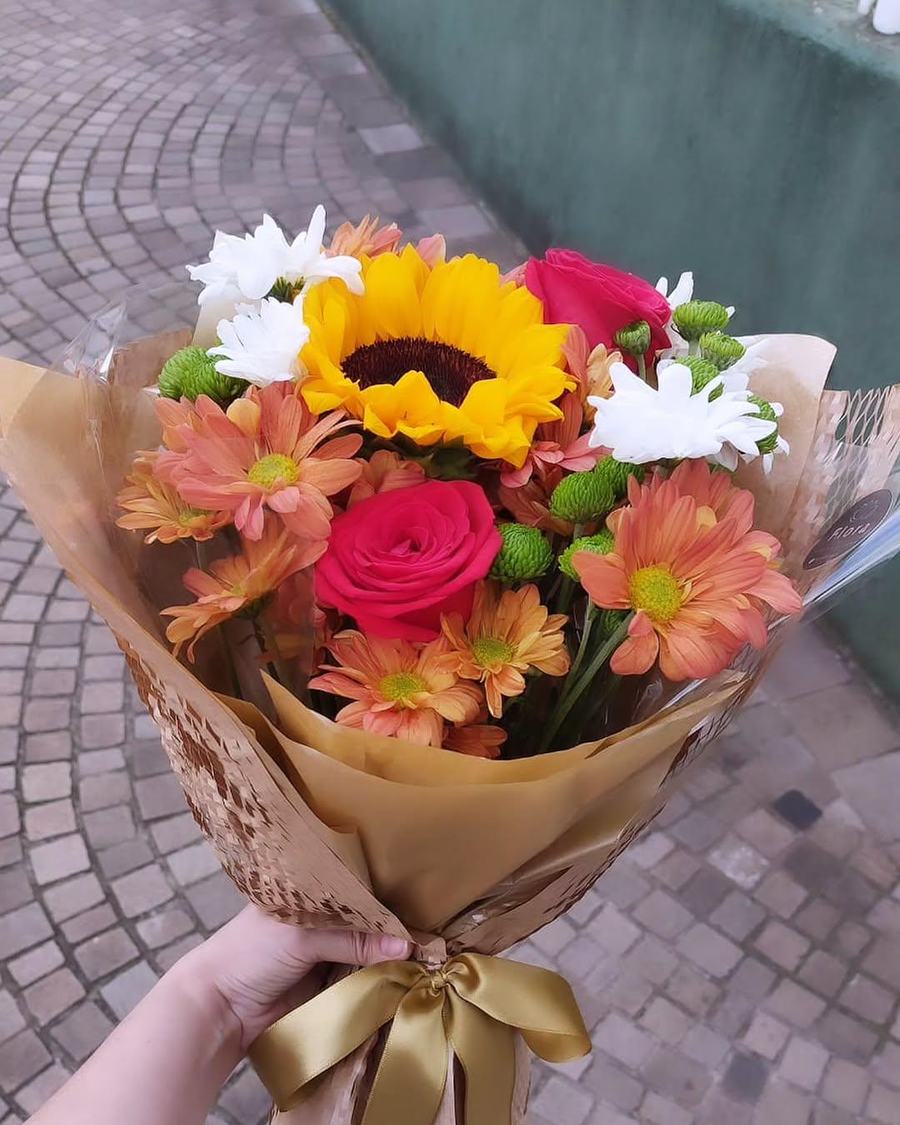 Buquê nº 03 — Tarde Dourada: Girassol, rosas pink e margaridas em tons de coral, embrulhados em kraft com laço de cetim dourado